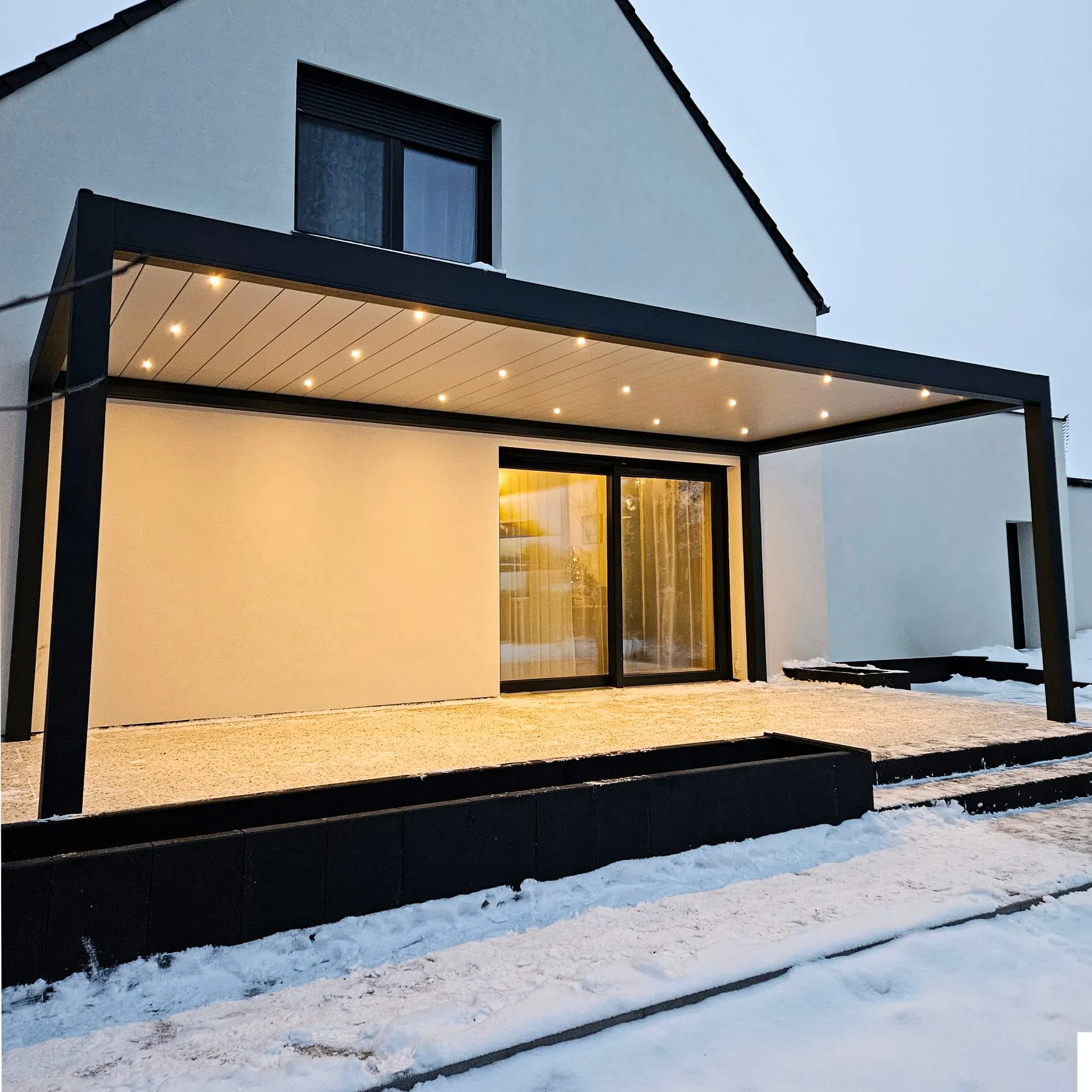 Wandmontierte Aluminium-Pergola mit geschlossenem Lamellendach und integrierten LED-Spots am Abend
